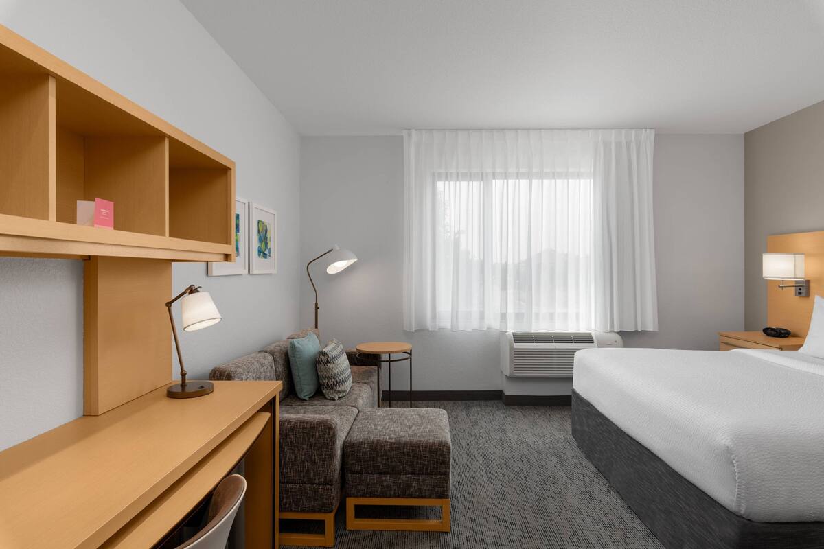 Una habitación de hotel con cama, escritorio con lámpara, sofá, una mesita y ventana con cortinas.