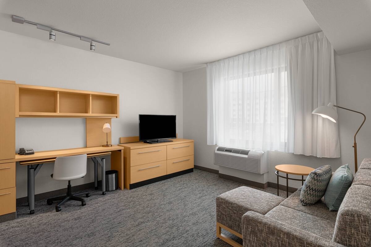 Una habitación de hotel moderna con un escritorio de madera, televisor, sofá y un amplio ventanal con cortinas.