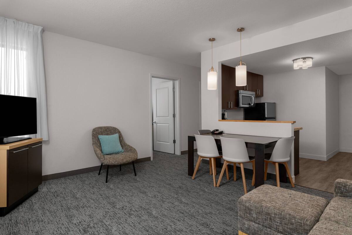 Una sala de estar moderna con un televisor de pantalla plana, un sofá, un comedor con una mesa y sillas, una cocina con microondas y una puerta que da a otra habitación.
