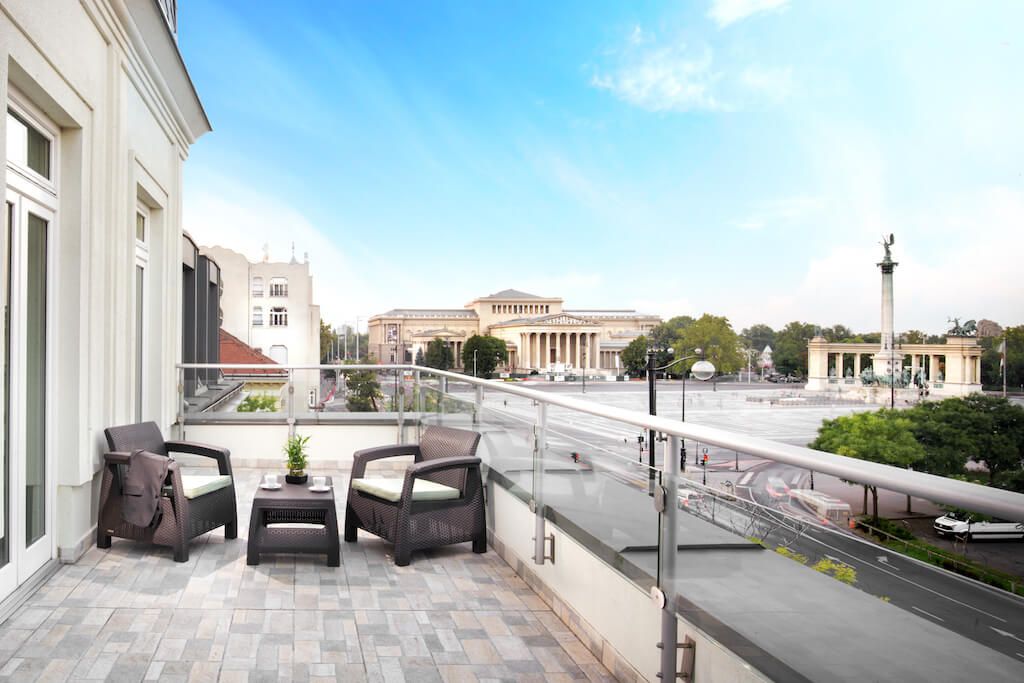 A balcony with two wicker chairs and a small table, overlooking a city street and a large building with classical architecture.