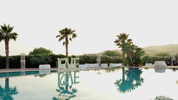 Una piscina con palmeras, un pabellón blanco y mesas dispuestas para un evento.