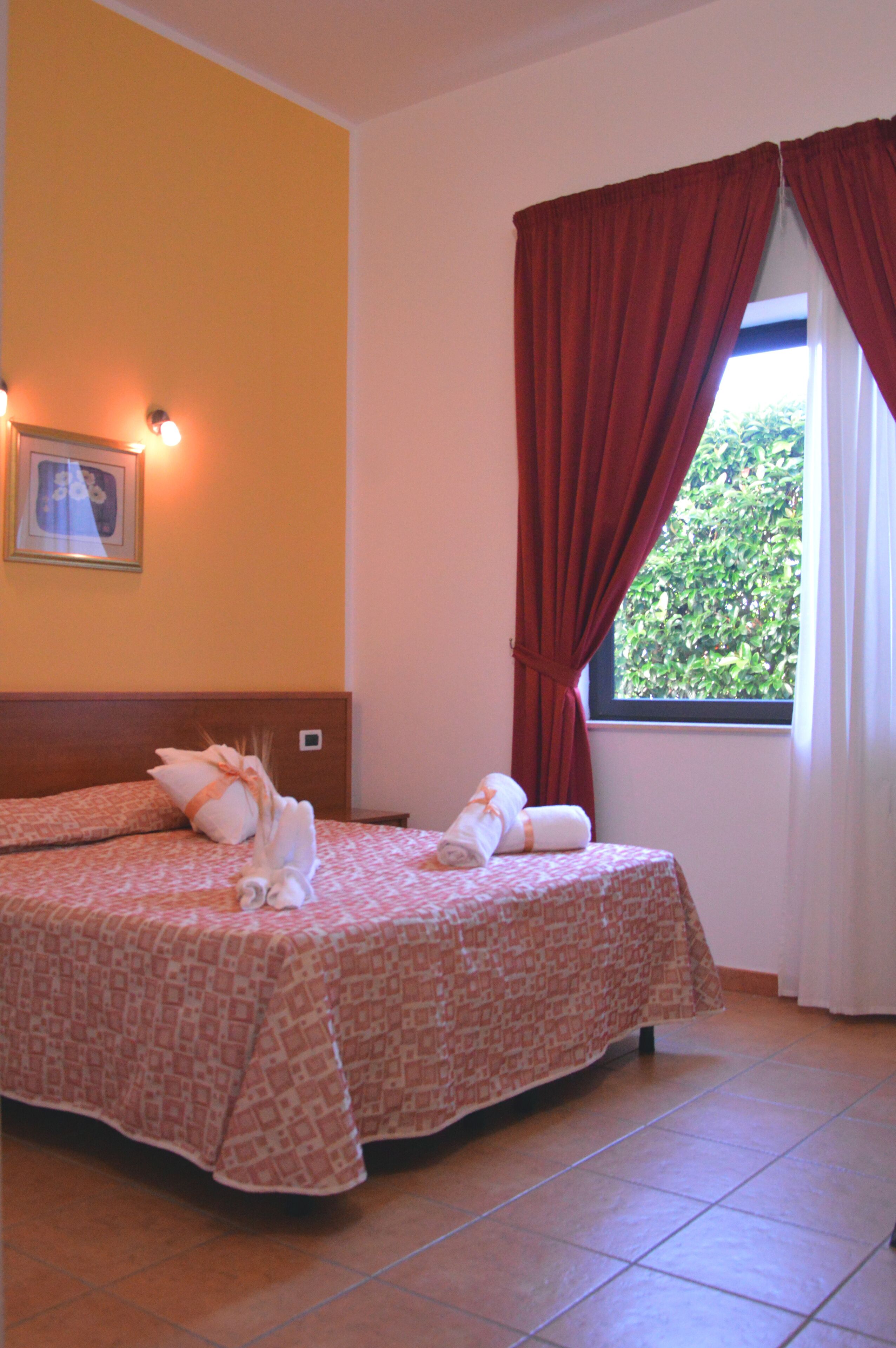 Habitación de hotel con dos camas, una ventana con cortinas rojas, un cuadro enmarcado en la pared y suelo de baldosas.