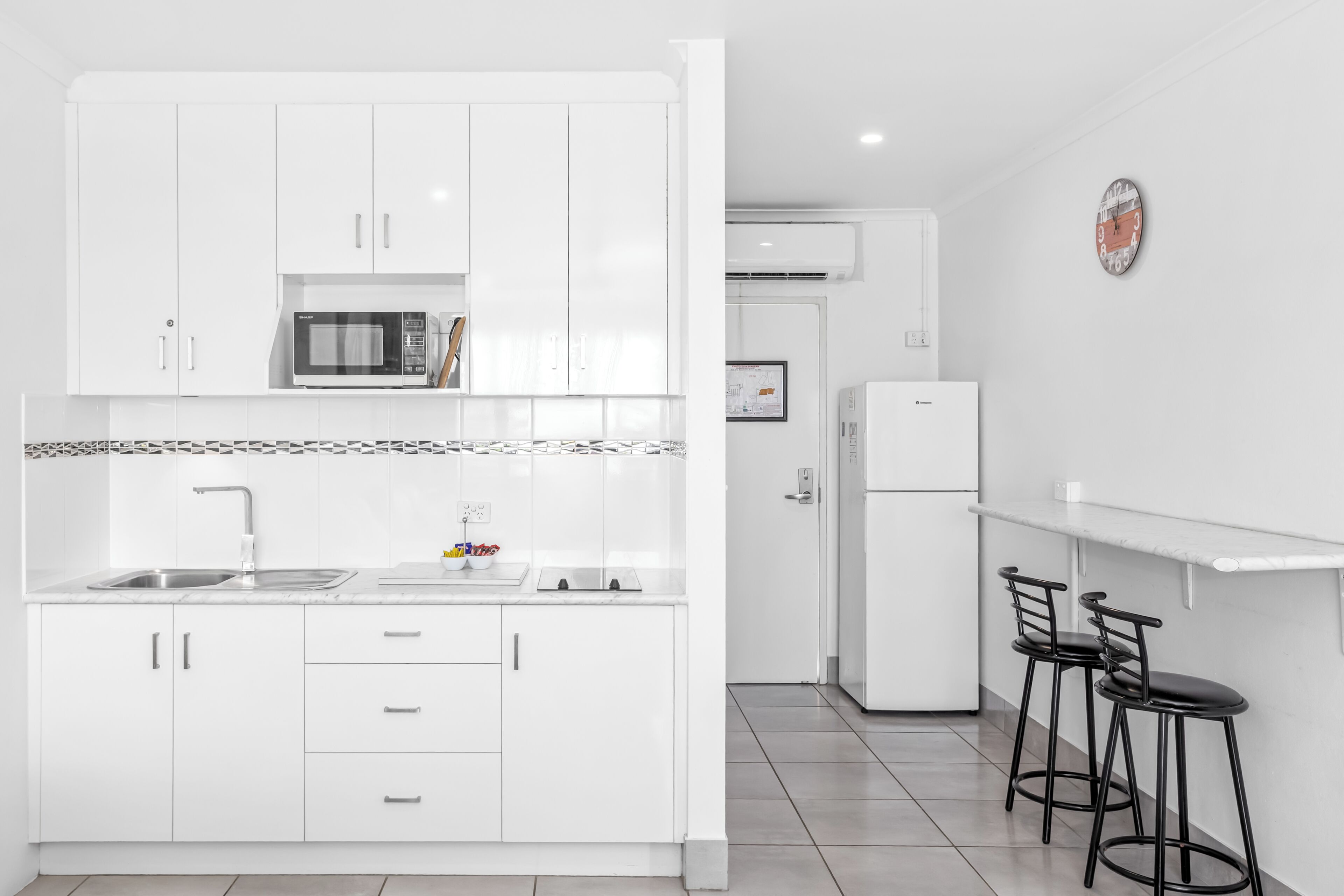 A modern kitchen with white cabinets, a microwave, a sink, and a refrigerator. There are two black bar stools and a white countertop.