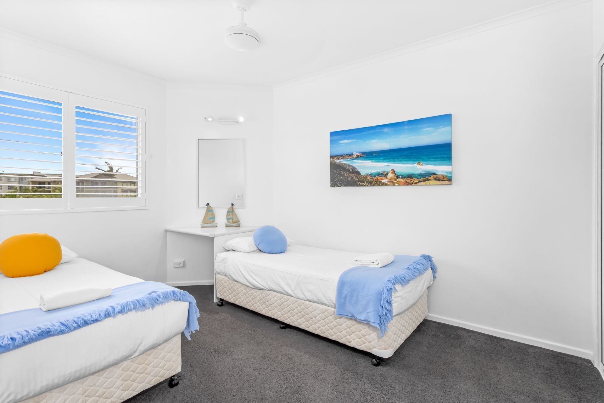 Een slaapkamer met twee bedden, een raam met zonwering, een schilderij van een strand en een plafondventilator.