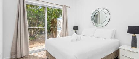 A bedroom with a large bed, two bedside tables, a round mirror, and a view of greenery through a sliding door.