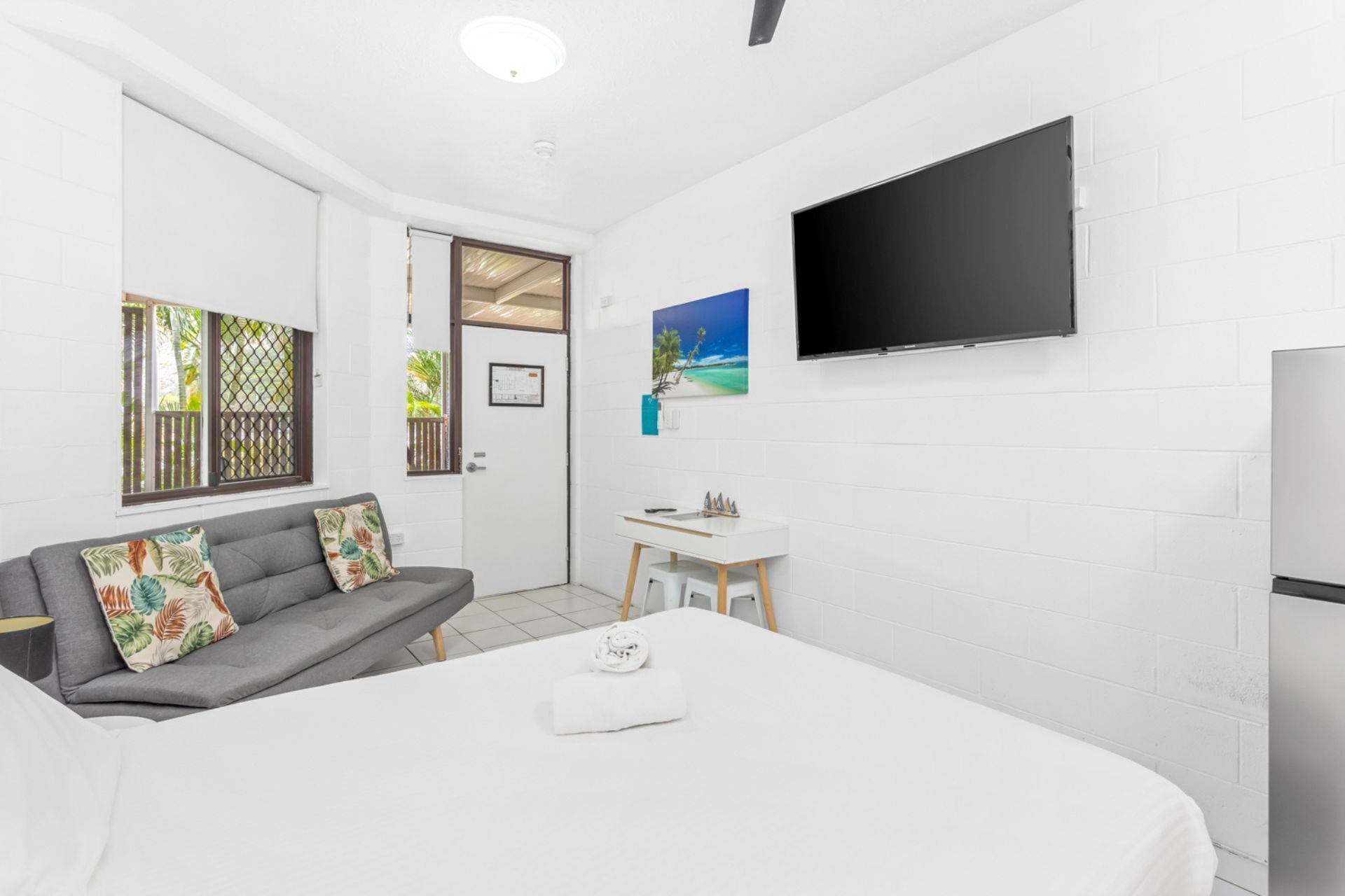 A hotel room with a bed, a gray sofa with floral cushions, a small white table, a TV mounted on the wall, and a door leading to a balcony.