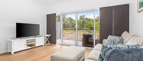 A modern living room with a flat-screen TV, a white entertainment unit, a grey sofa, and a large sliding glass door leading to an outdoor area.