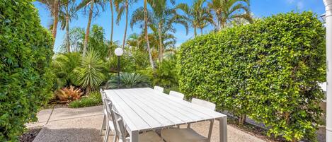 A white table and chairs set outdoors with a gravel path, surrounded by lush greenery and palm trees.