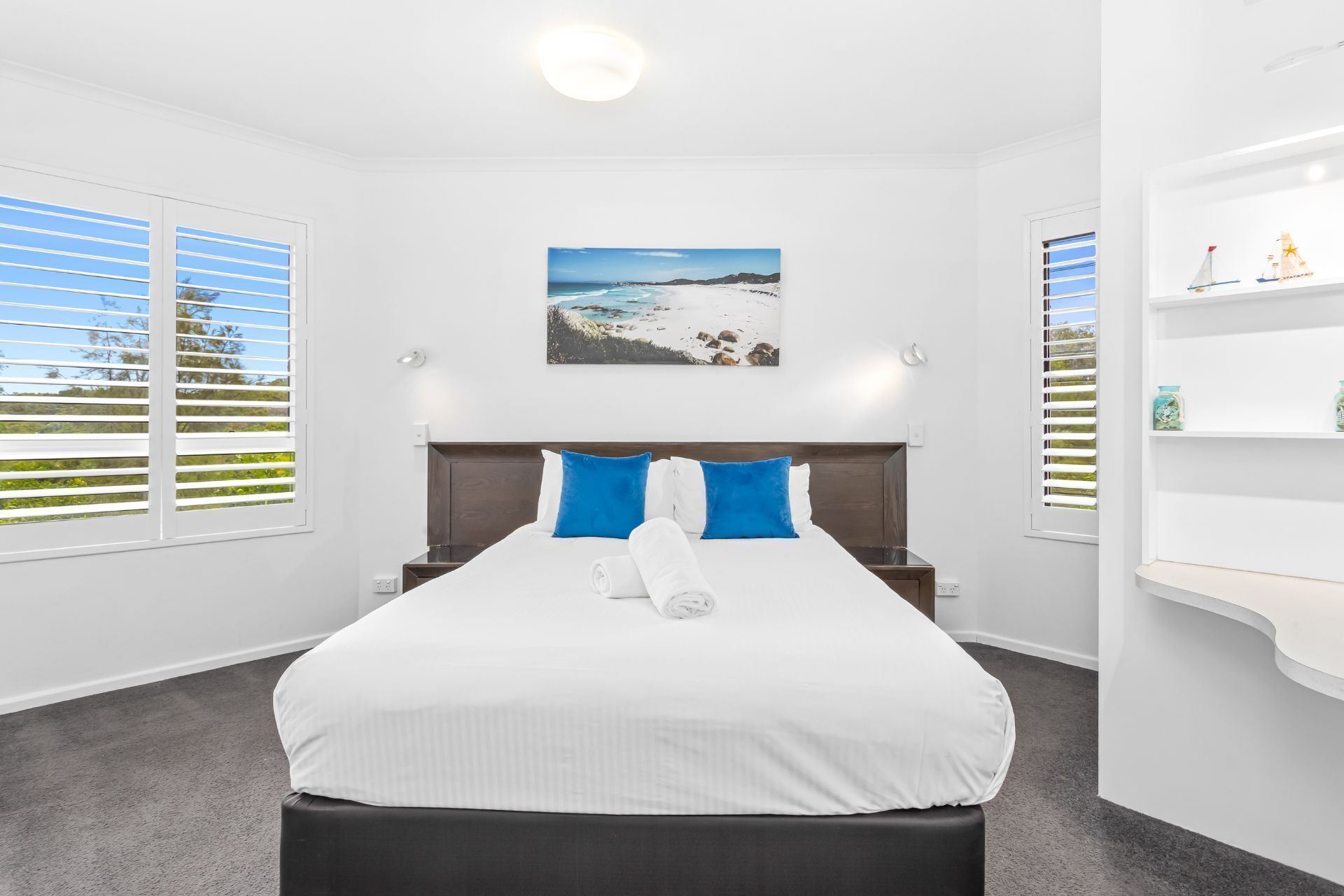 A hotel room with a bed, a painting of a beach, a nightstand, and a shelf with items.