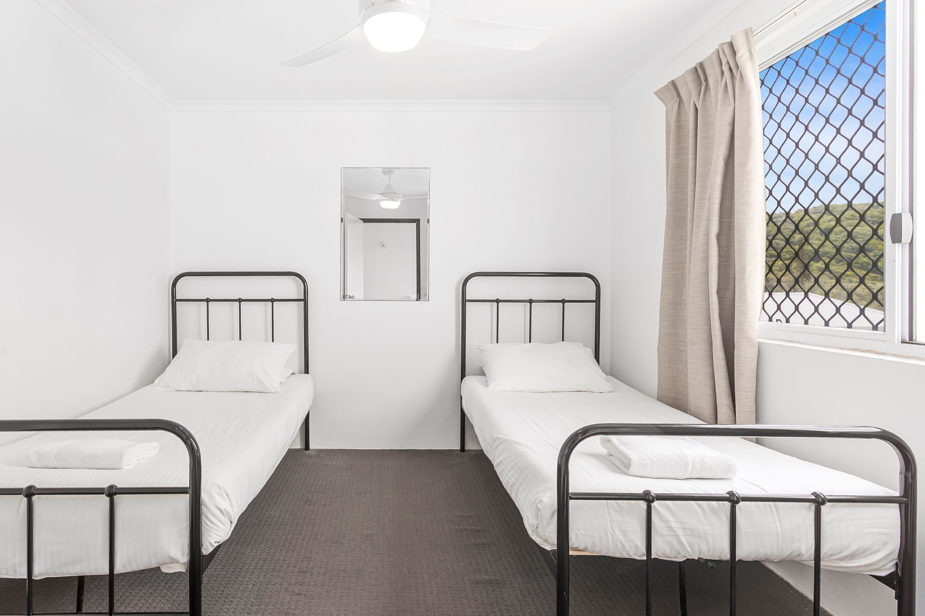 A room with two single beds, a ceiling fan, a window with curtains, and a mirror on the wall.