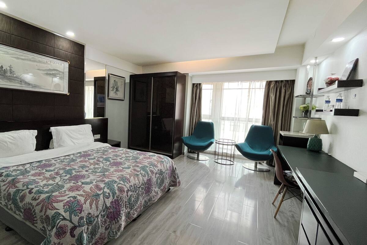 A hotel room with a large bed, two chairs, a desk, a wardrobe, and a window with curtains.