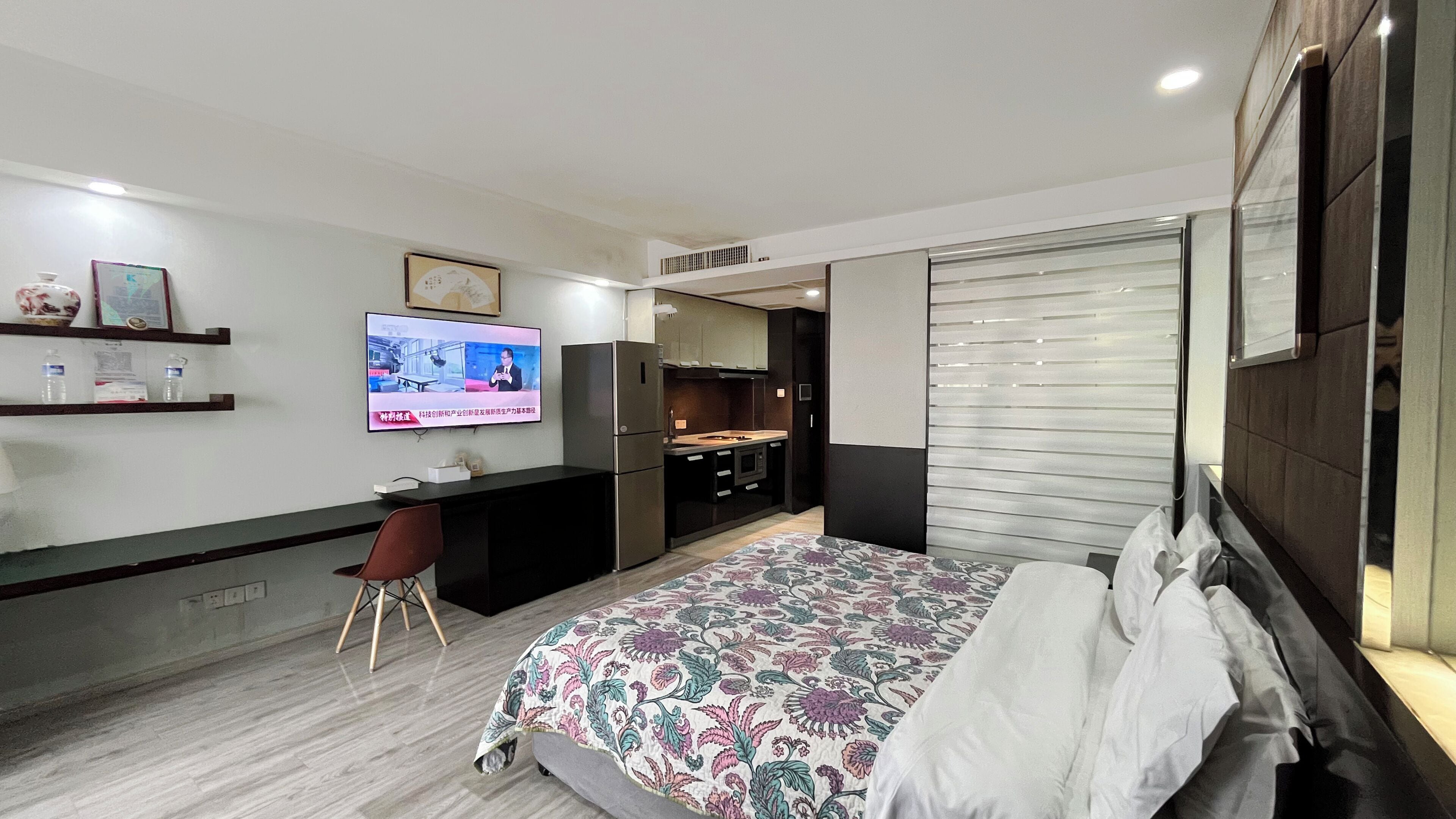 A modern hotel room with a bed, a desk, a TV, and a kitchenette.