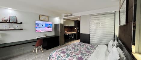 A modern hotel room with a bed, a desk, a TV, and a kitchenette.