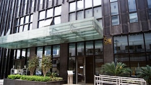 A modern building with a glass entrance canopy, a sign indicating the name and address, and a landscaped area with plants and a metal railing.