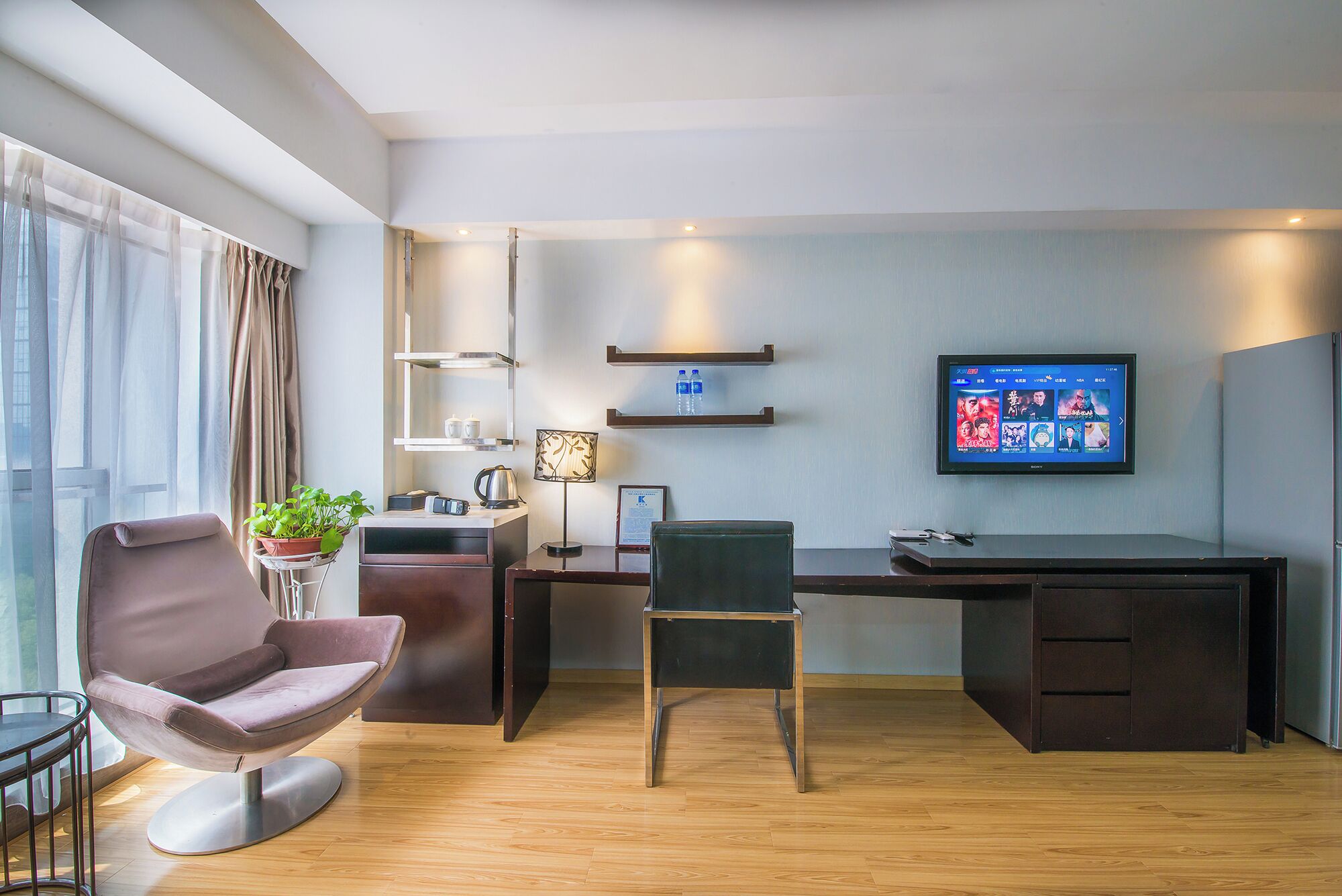A modern hotel room with a wooden desk, a comfortable chair, a flat-screen TV, a small table with a plant, and a window with sheer curtains.