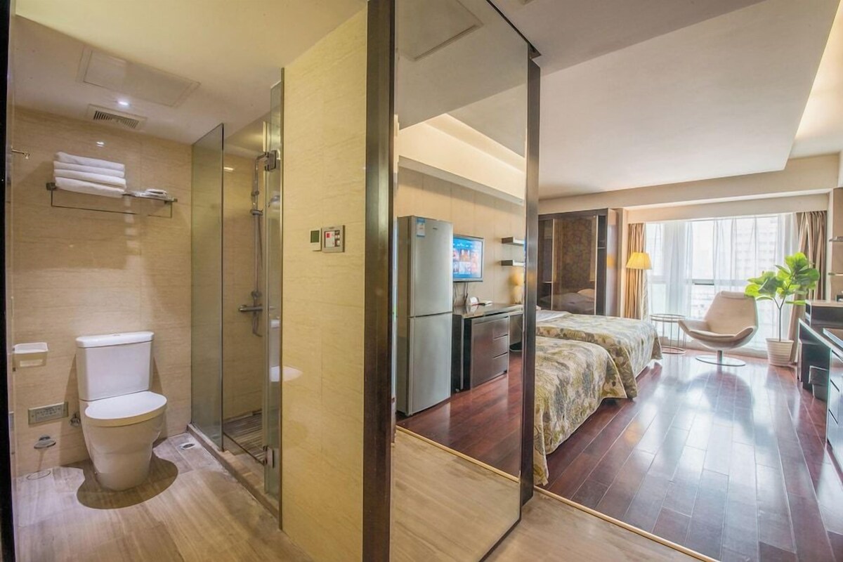 A hotel room with a glass shower enclosure, a bed with a floral bedspread, a television, and a desk with a chair.