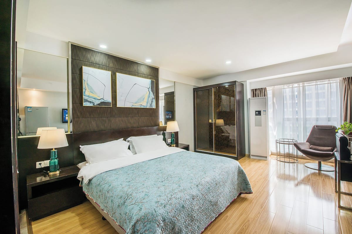 Une chambre d’hôtel moderne dotée d’un grand lit, d’un coin salon avec un fauteuil et une petite table, d’un minibar et d’une grande fenêtre avec des rideaux.