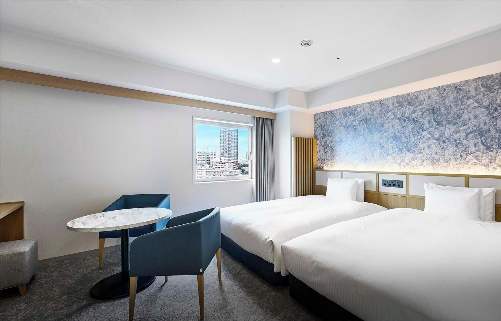 Habitación de hotel con dos camas, una mesita redonda con sillas, ventana con vistas a la ciudad y una pared con diseño de mármol.