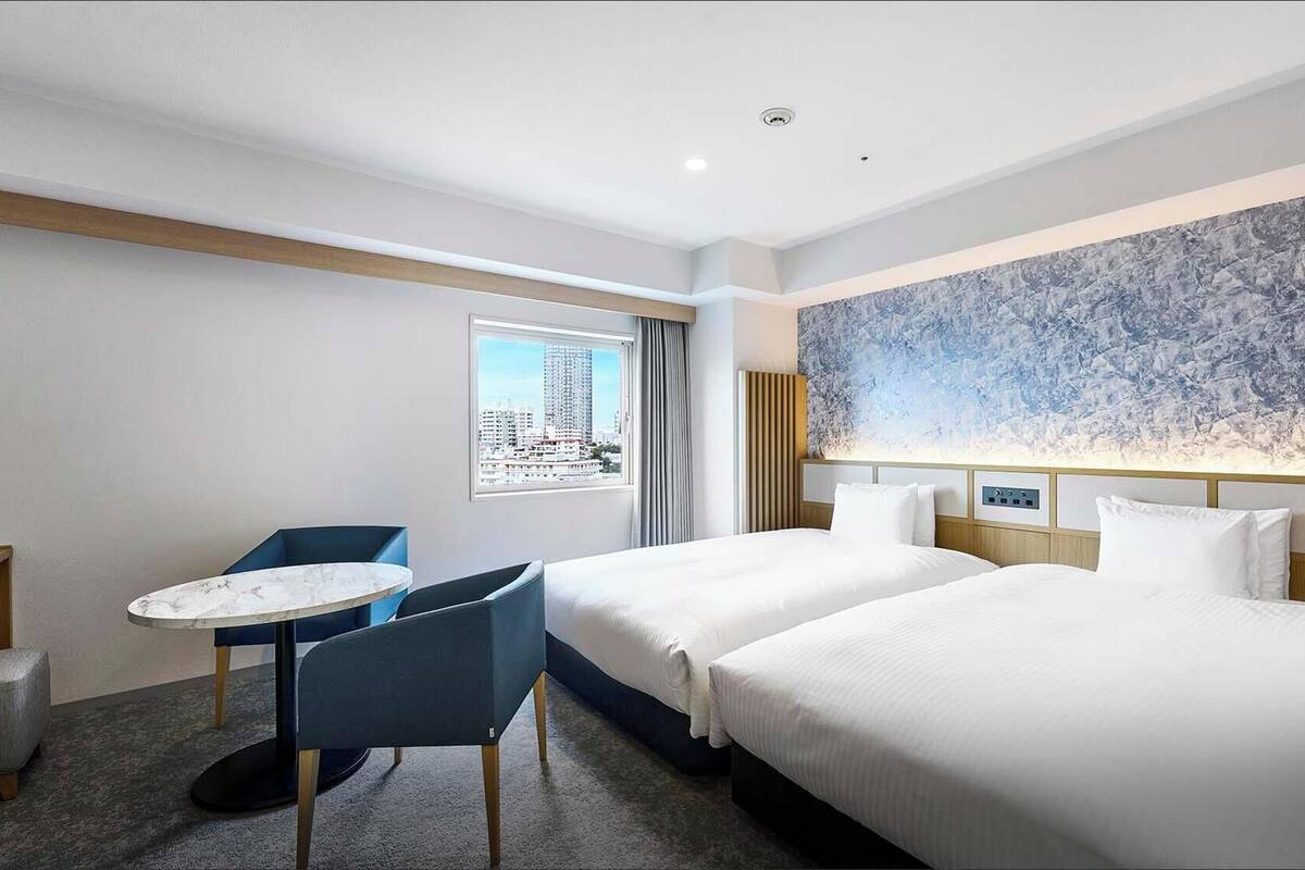 A hotel room with two beds, a small round table with chairs, a city view window, and a marble-patterned wall.