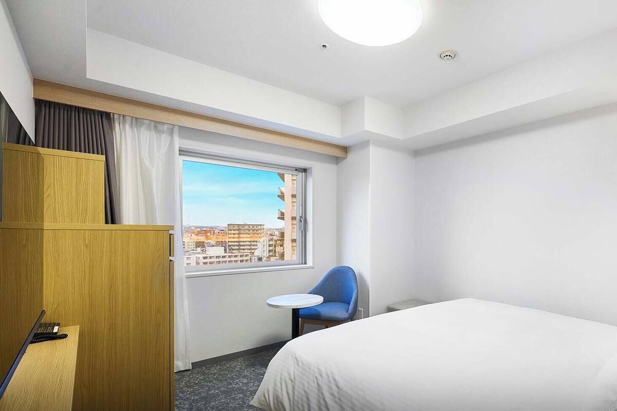 A hotel room with a bed, a desk, a chair, and a window with a city view.