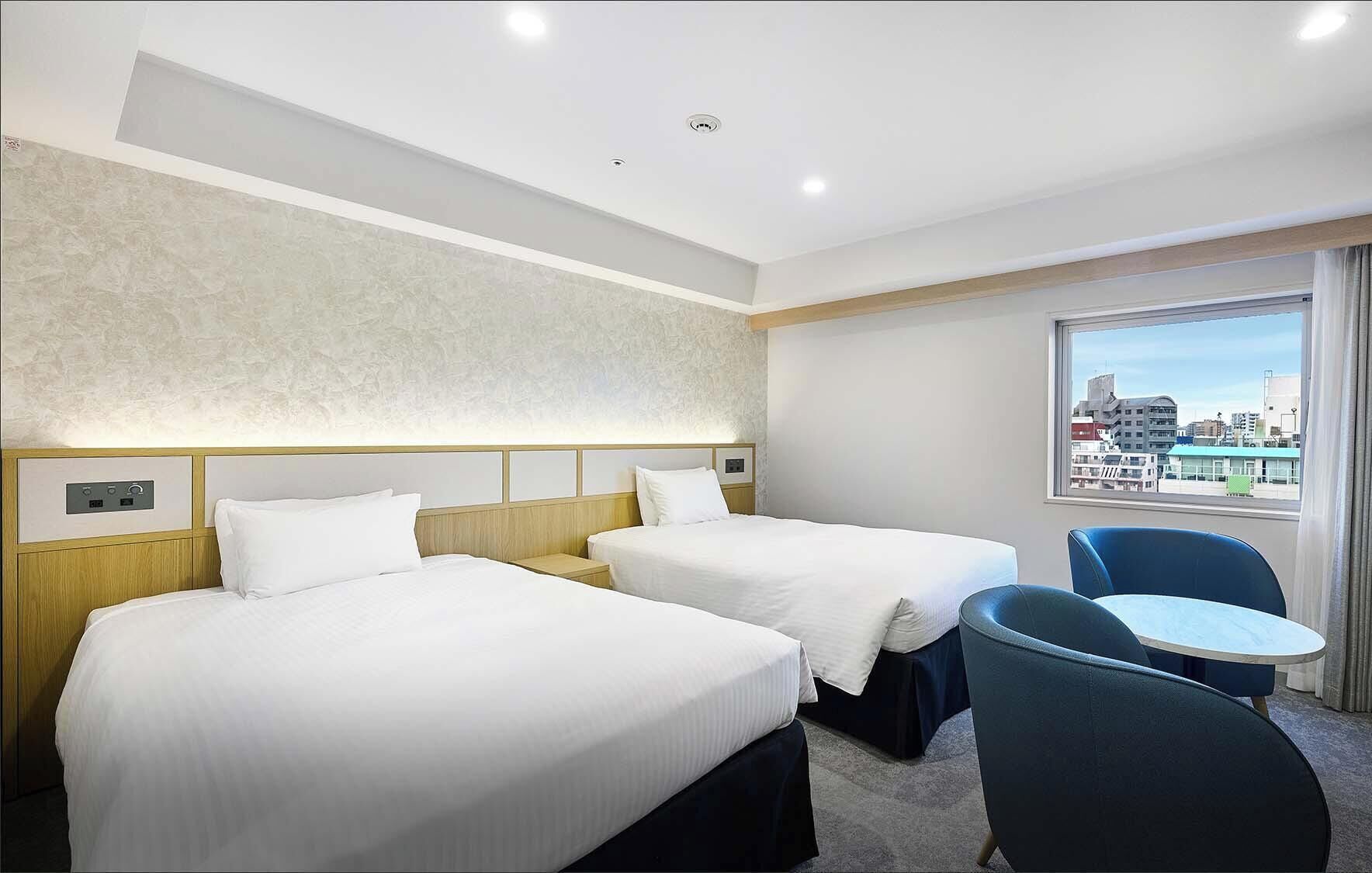A hotel room with two beds, a small table, a chair, and a window with a city view.