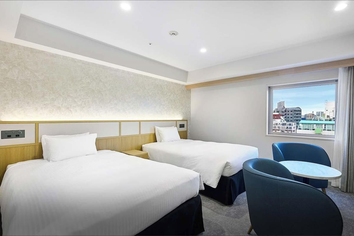 Habitación de hotel con dos camas, una mesita, una silla y ventana con vista a la ciudad.