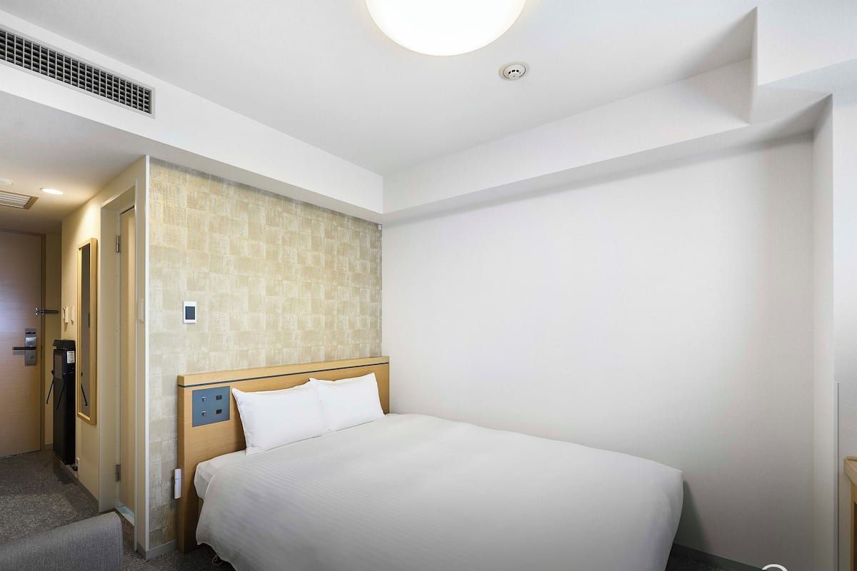 A hotel room with a bed, a wooden headboard, a small bedside table, a wall-mounted TV, and a door leading to a bathroom.