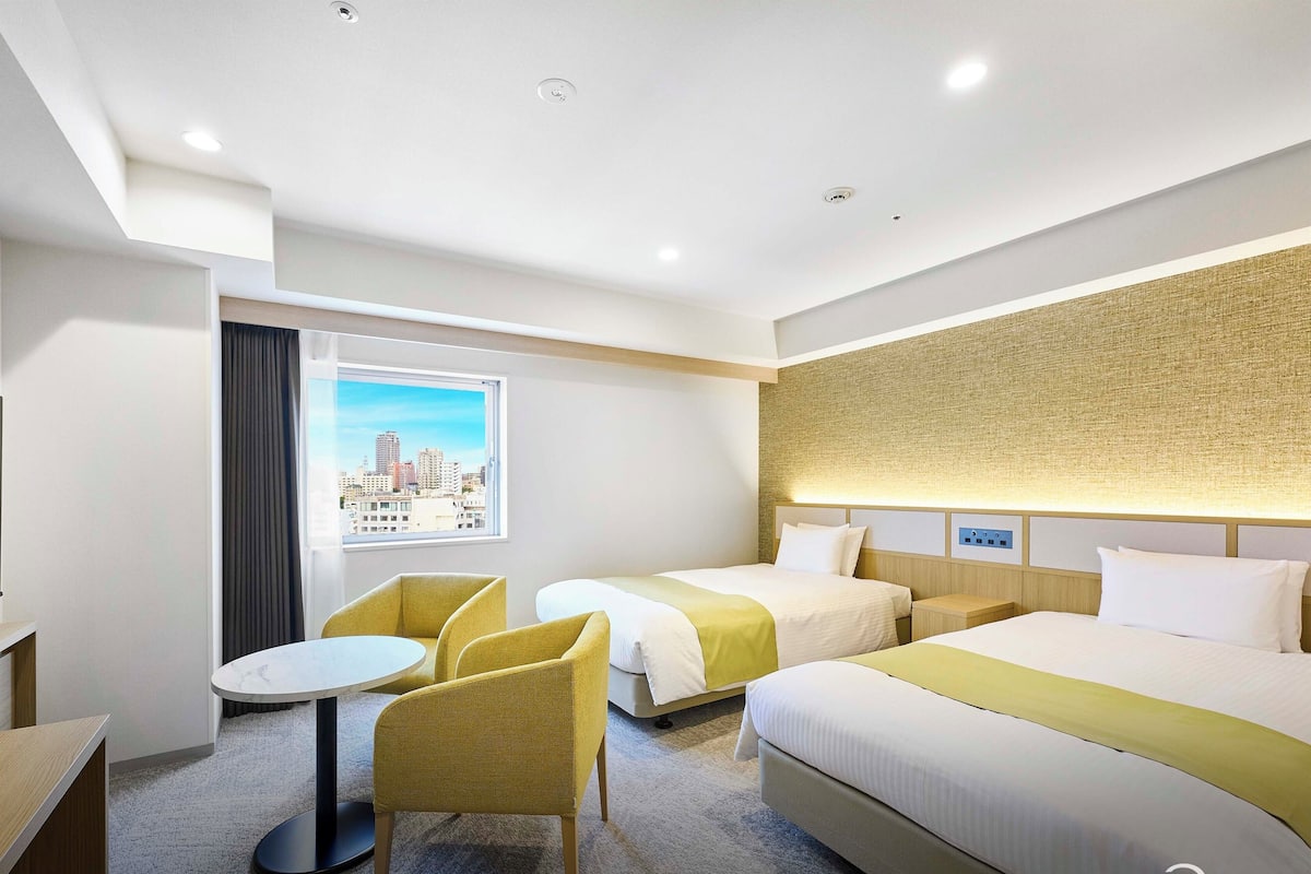 A hotel room with two beds, a small table, and chairs. There is a window with a city view.