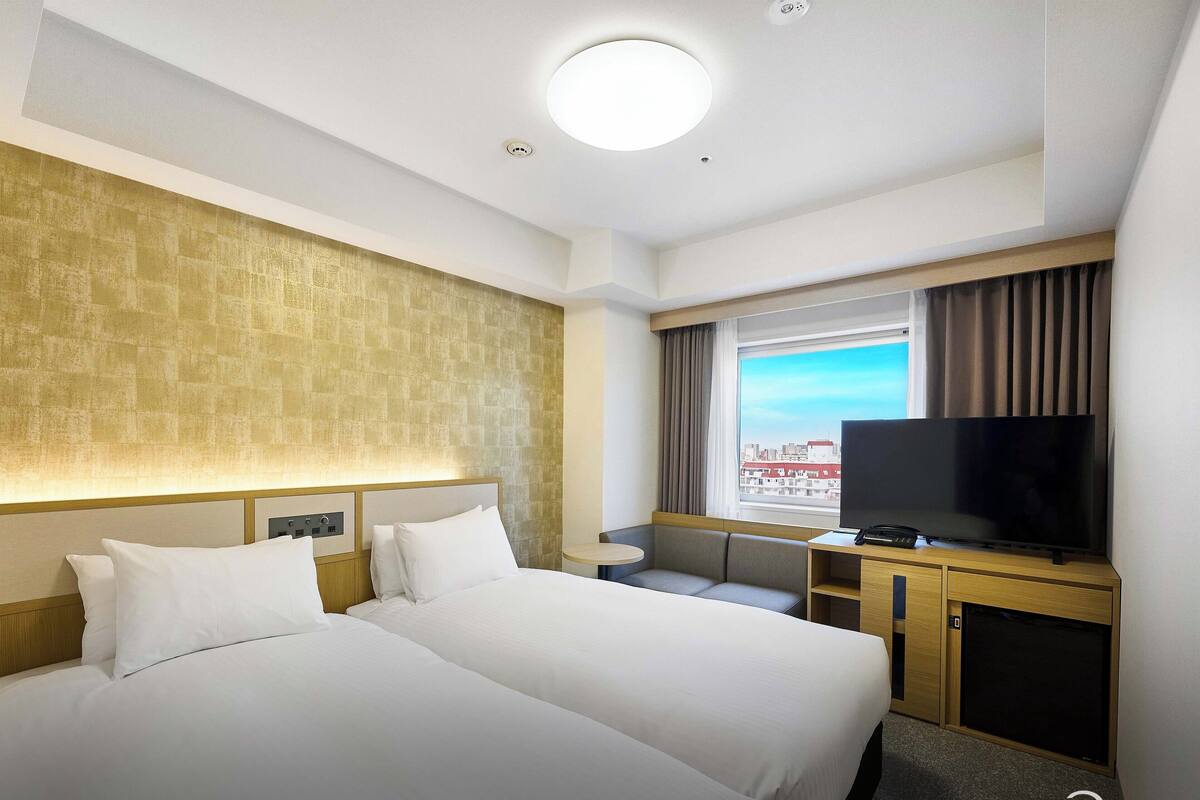 A hotel room with two beds, a TV, a small table, and a window with a city view.