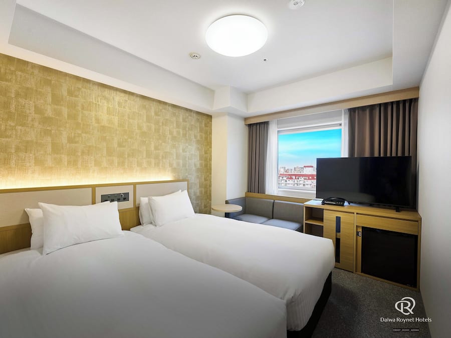Habitación de hotel con dos camas, televisor, una mesita y ventana con vista a la ciudad.