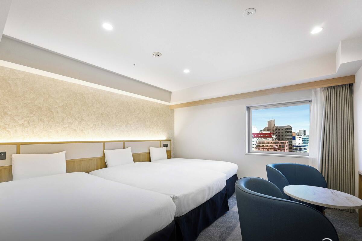 A hotel room with two beds, a small table, a chair, and a window with a city view.