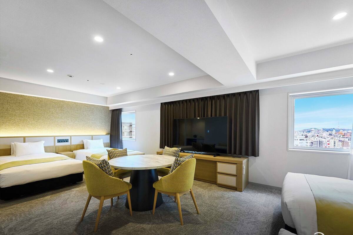 A modern hotel room with a large bed, a dining table with chairs, a TV, and a city view.