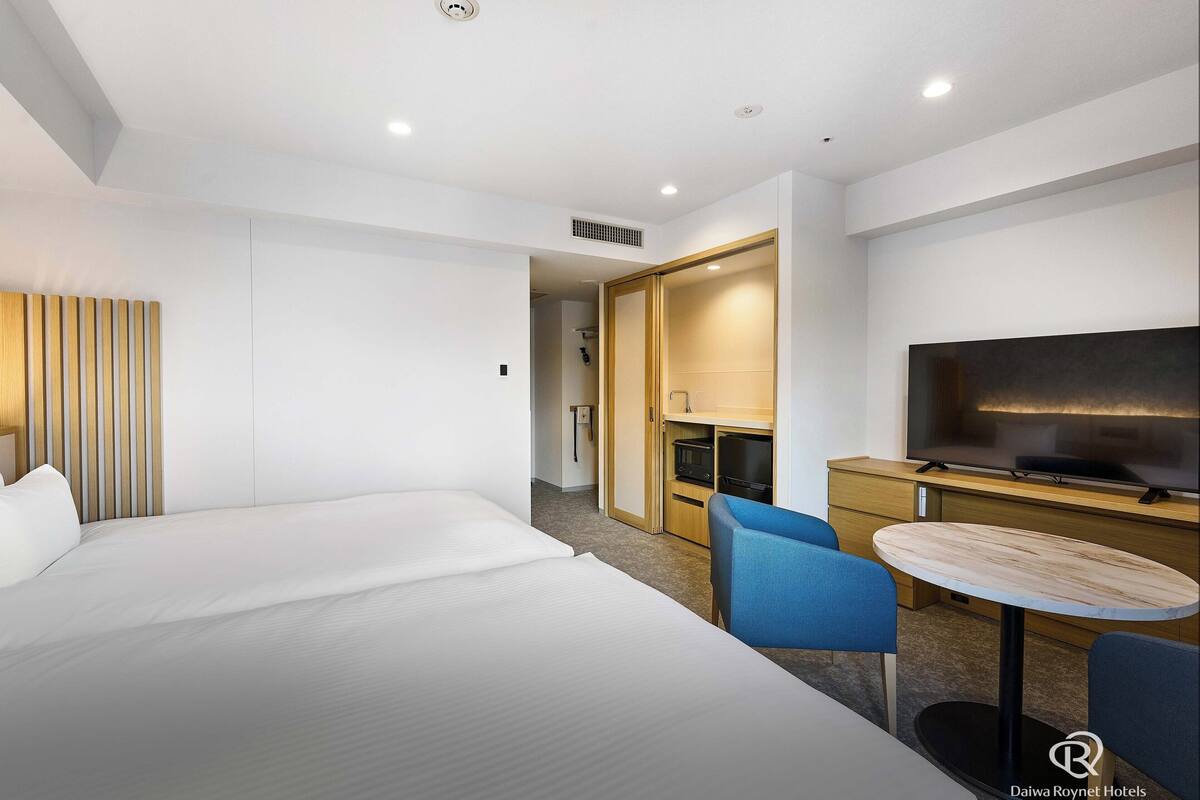 A hotel room with a large bed, a desk with a flat-screen TV, a chair, and a small table.