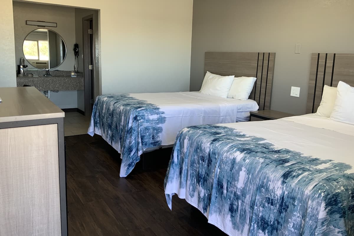A hotel room with two beds, a wooden desk, a bathroom with a mirror, and a view of the outdoors.
