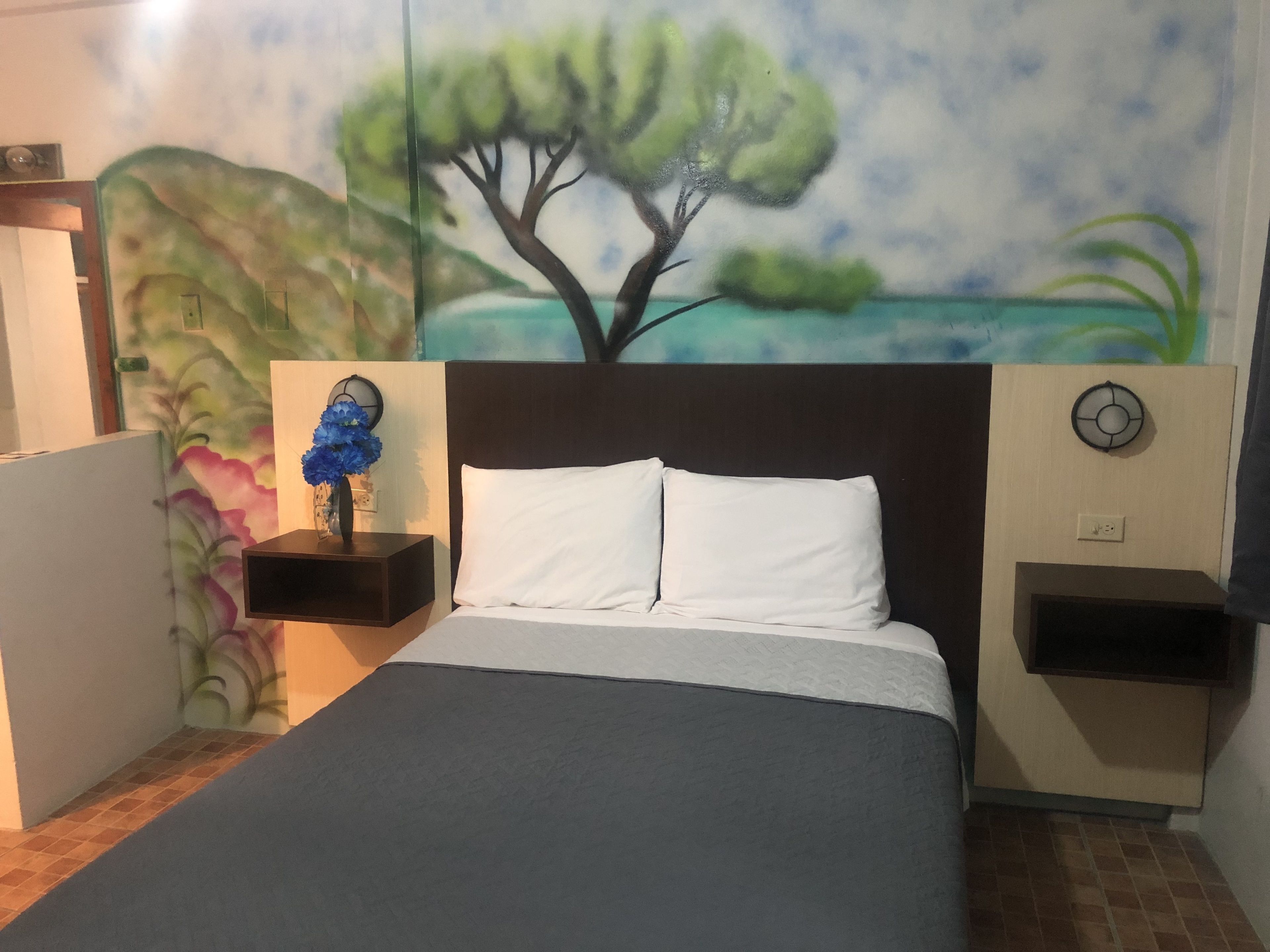 Un dormitorio con una cama, una mesita de noche con un florero y un reloj de pared, además de un cuadro de un árbol.