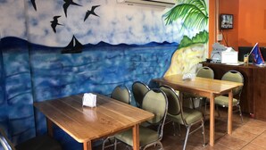 Ein Restaurant mit Wandgemälde von fliegenden Vögeln und einer Strandansicht. Es gibt Holztische und -stühle, eine Klimaanlage und einen Fernseher an der Wand.