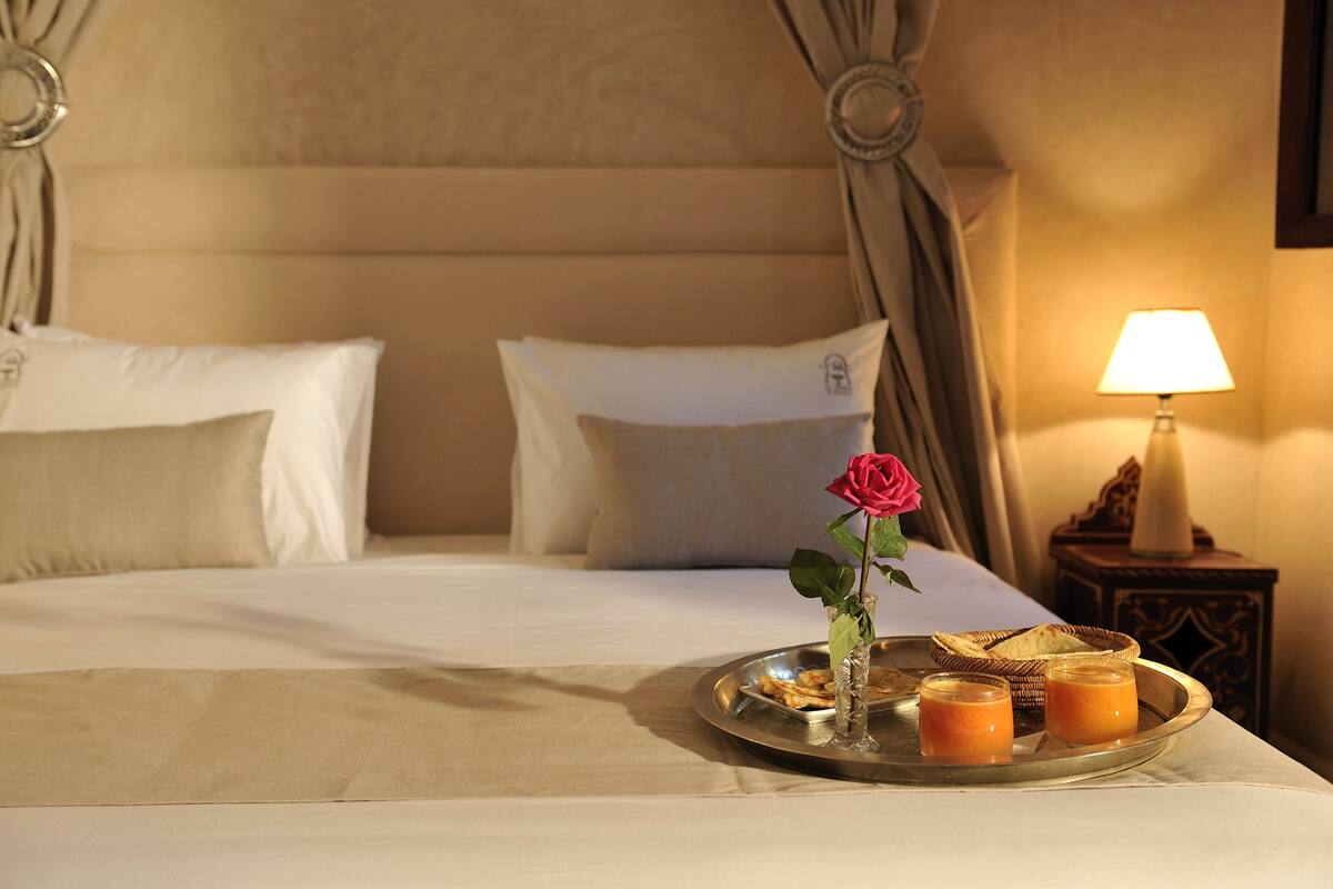 Un lit bien fait, avec du linge de lit blanc, un plateau avec de la nourriture et des boissons, et une rose posée sur le lit.