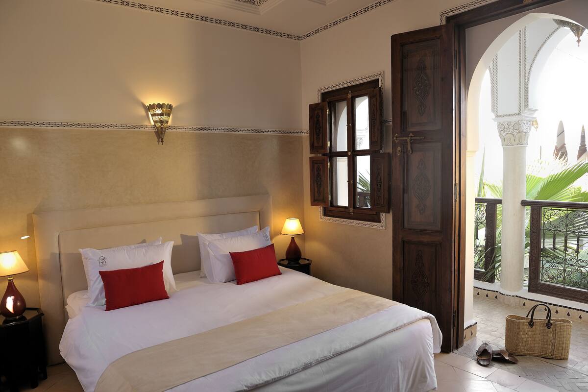 Une chambre d’hôtel avec un grand lit, deux lampes de chevet, une porte en bois et une vue sur une cour intérieure.