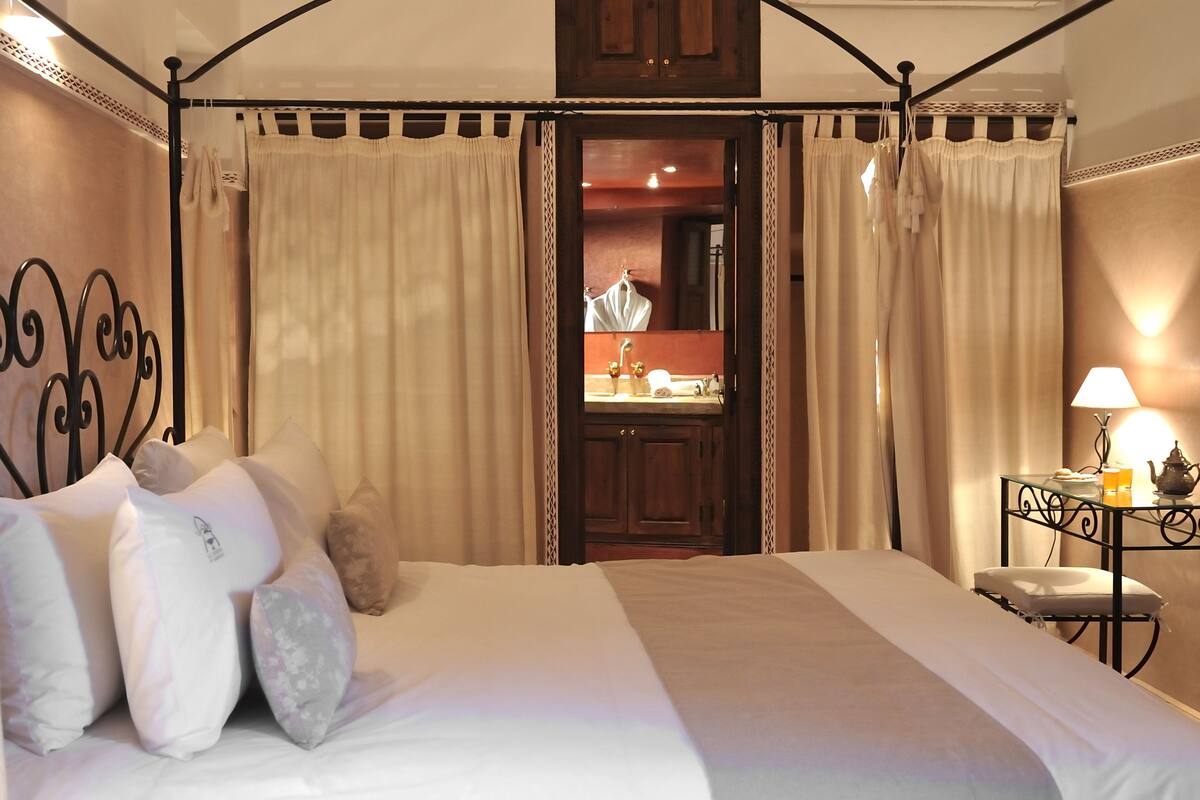 Un lit à baldaquin, une table de chevet avec une lampe et un miroir qui reflète la salle de bain.