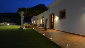 Un área de comedor al aire libre con techo, mesas y sillas, una pérgola con follaje, y una montaña al fondo.