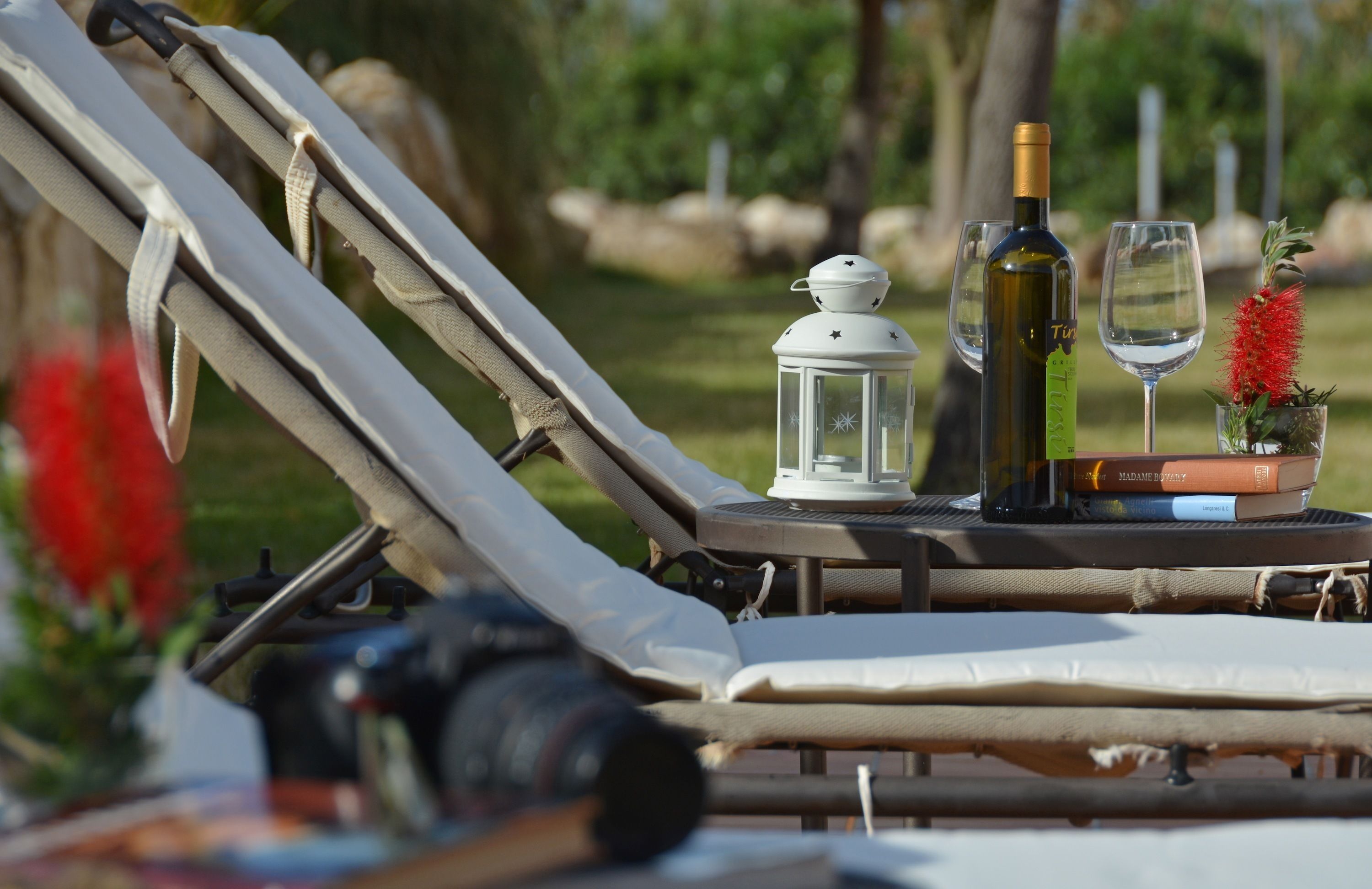 Una tumbona con una cámara, una botella de vino, una copa, un farol y libros sobre una mesa.