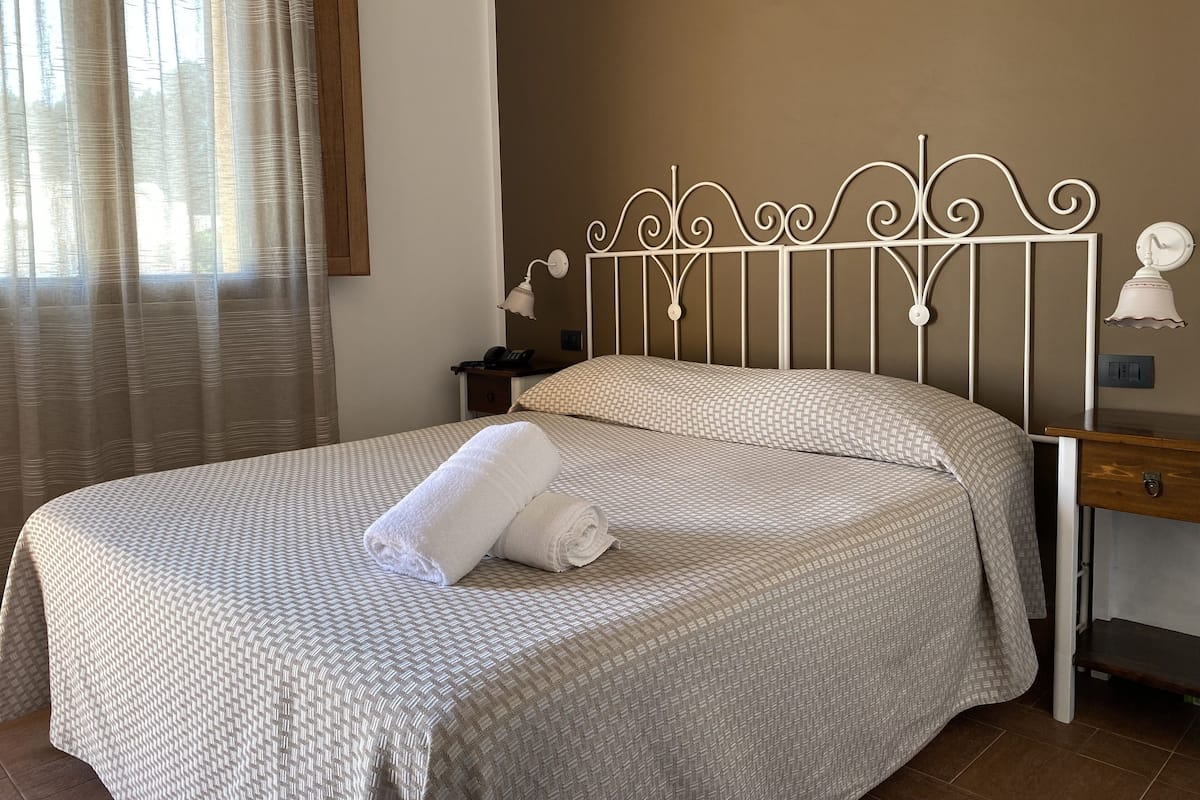 Una cama bien tendida con una colcha estampada y dos almohadas, una mesita de noche con una lámpara, y una ventana con cortinas.
