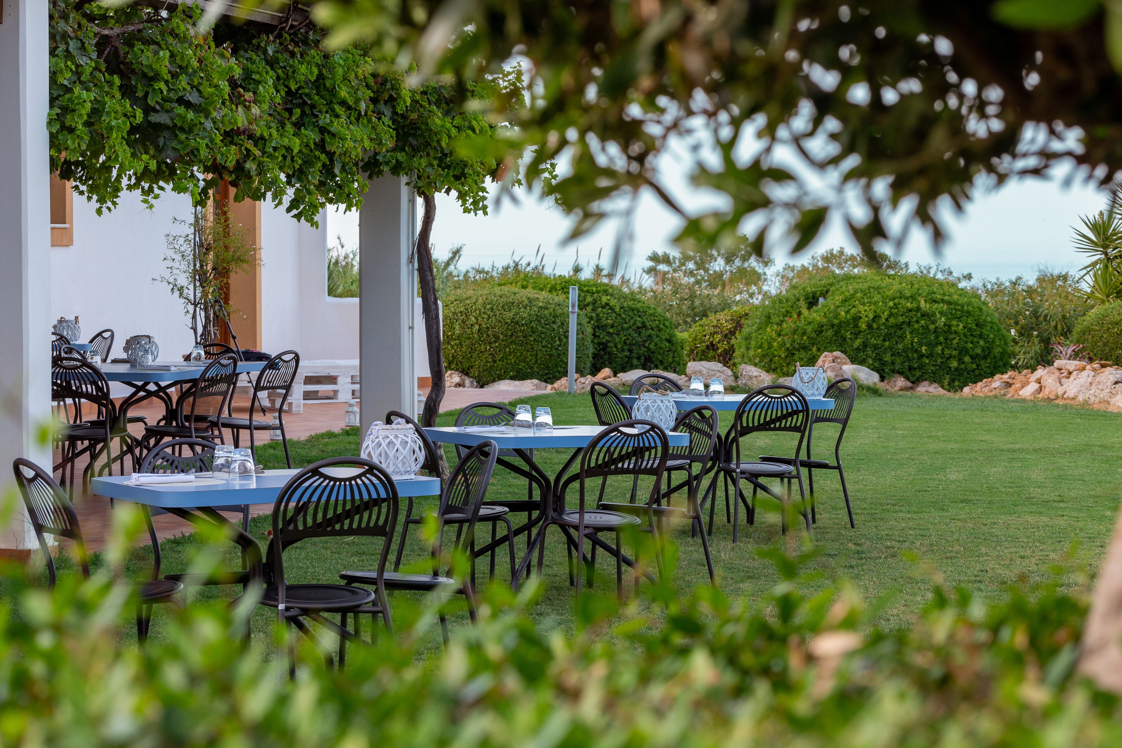 An outdoor dining area with tables and chairs, surrounded by greenery and a view of the sea.