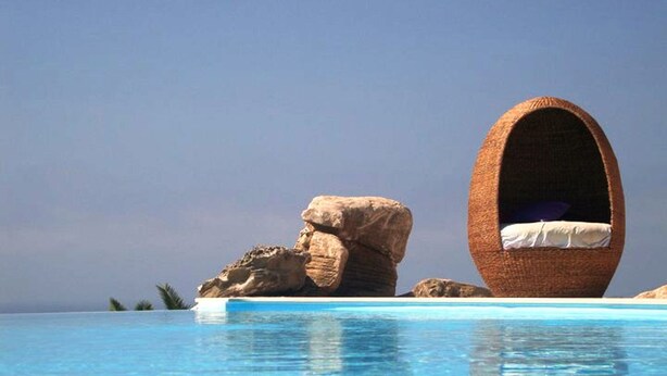 A clear blue pool with a wicker chair and a rock formation.