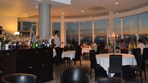 A restaurant with round tables, a bar area, and large windows offering a city view.