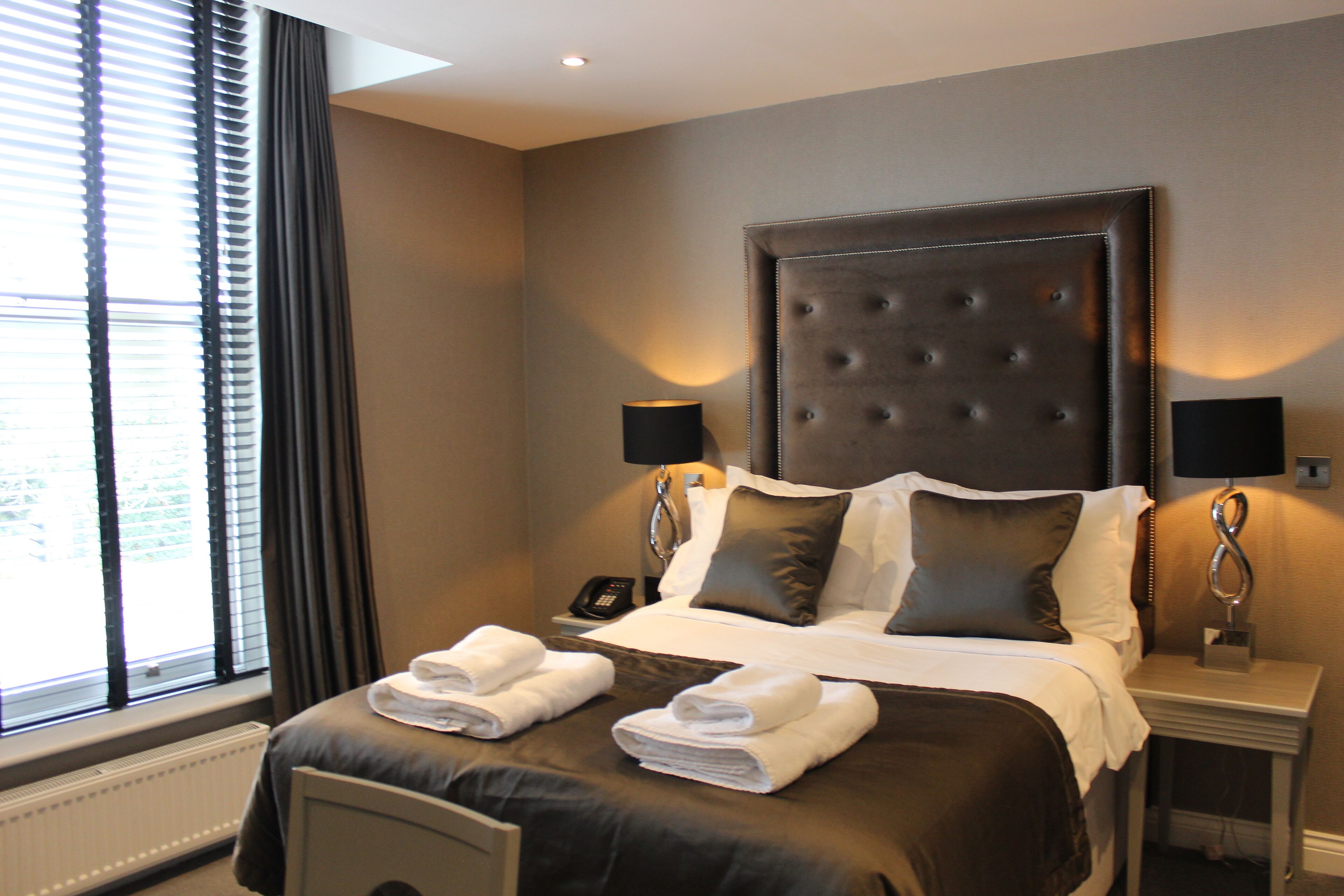 A hotel room with a large bed, two bedside lamps, a nightstand with a phone, and a window with blinds.