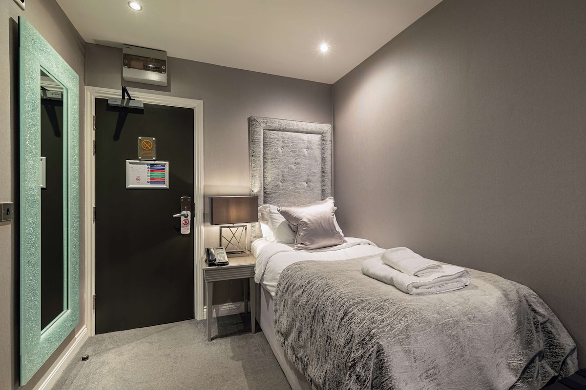 A hotel room with a bed, a bedside table with a lamp, a door with a security card reader, and a mirror.