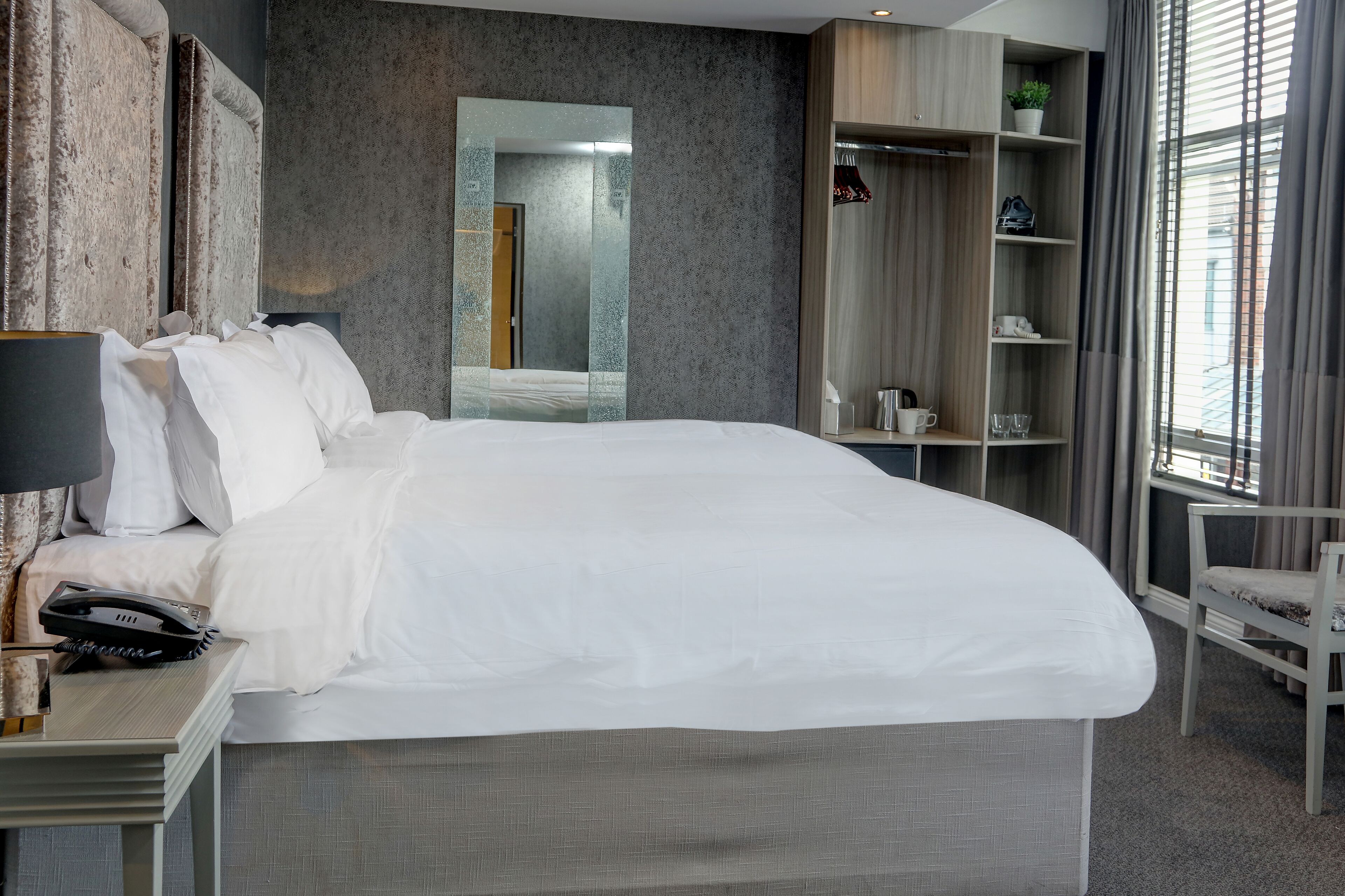 Une chambre d’hôtel moderne dotée d’un grand lit, d’un téléphone sur la table de chevet, d’un miroir, d’une armoire et d’une fenêtre avec des rideaux.