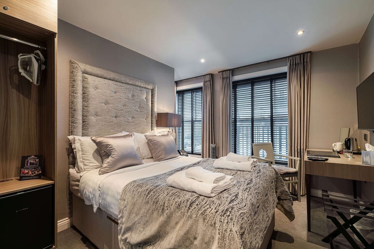 A hotel room with a large bed, bedside table, desk, and a window with blinds.