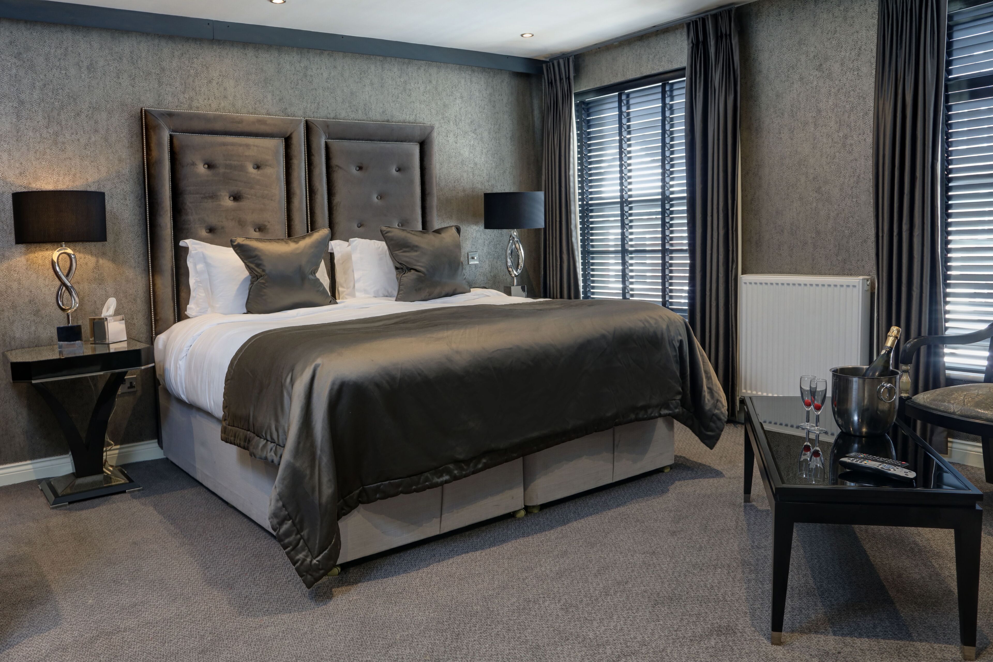 A modern hotel room with a large bed, a bedside table with a lamp, a small table with a bottle and glasses, and a chair.